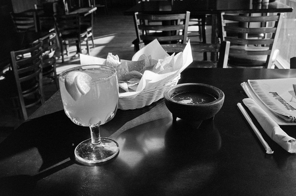 Black and white photo of a tabletop containing margarita glass with a bowl of tortilla chips and salsa.