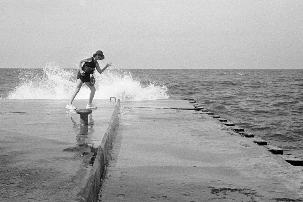 Black and white photo of woman waving on a pier with wave crashing behind her