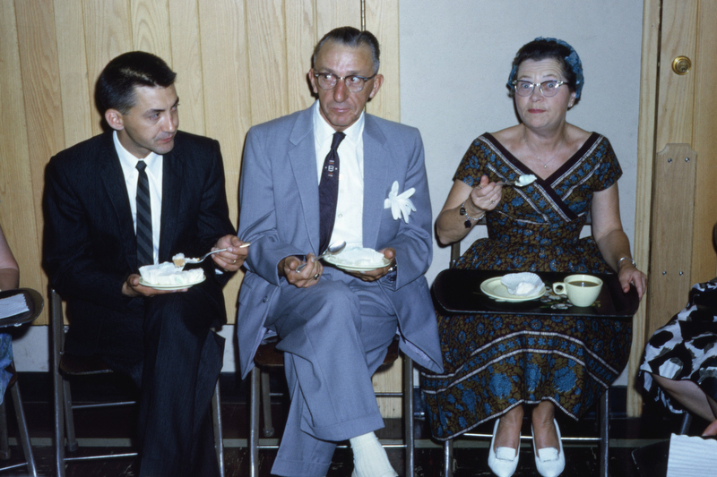 People in chairs, eating, at a reception. 1960s.
